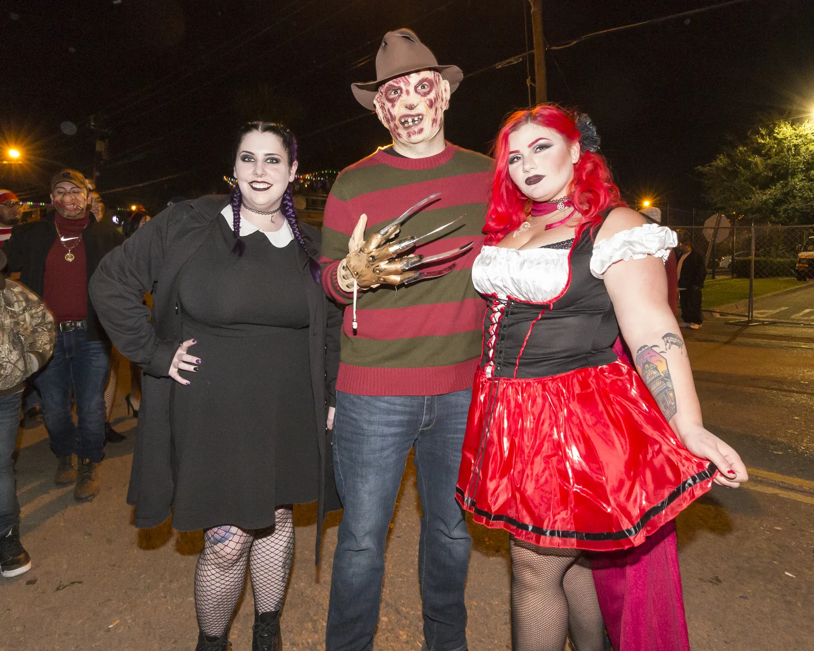 Three adults in Halloween costumes on a street at night: a woman dressed in black like Wednesday Addams, a man as Freddy Krueger with a striped sweater and claw glove, and a woman in a red dress as Little Red Riding Hood.