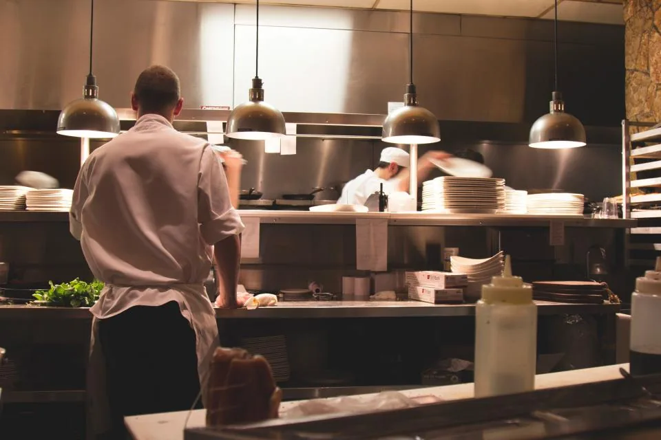 Two cooks in white jackets work the pass of a busy restaurant kitchen under hanging lamps, with stacks of clean plates waiting on the shelf.