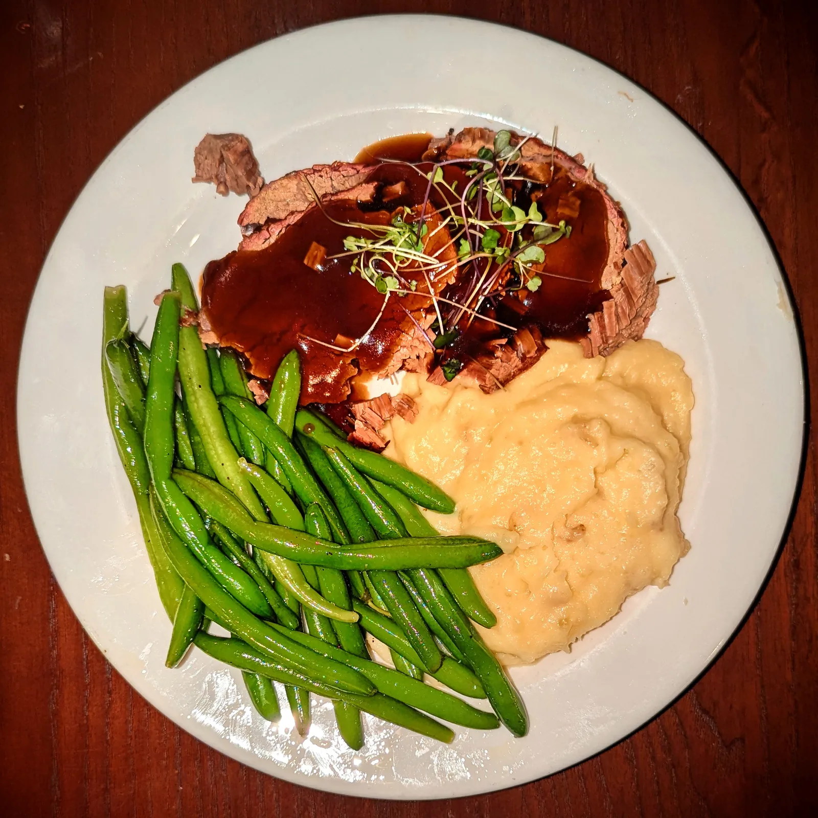 Sliced pot roast in dark gravy on a white plate, next to a scoop of mashed potatoes and a pile of green beans.